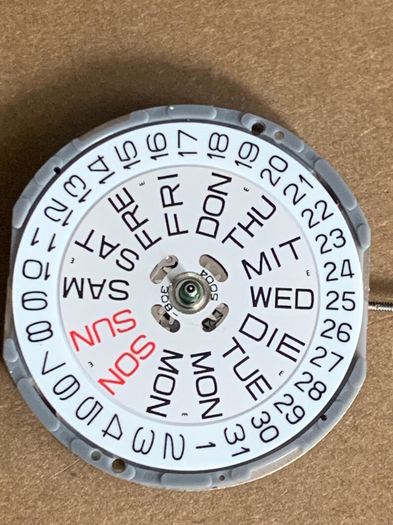 May include: A watch calendar wheel with a white face, featuring days of the week in English and numbers 1-31. The day "SUN" is in red. The wheel is surrounded by a gray ring.