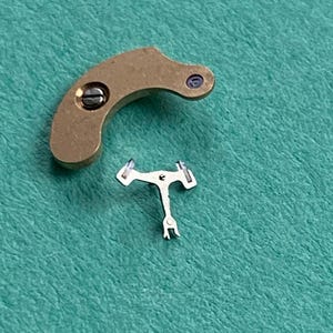 May include: Two small watch parts. One is a curved, brass-colored piece with a screw and a small purple dot. The other is a small, silver-colored, T-shaped piece with two arms and a small fork at the bottom.