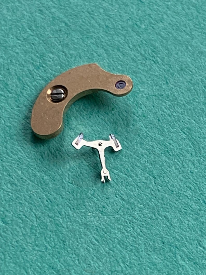 May include: Two small watch parts. One is a curved, brass-colored piece with a screw and a small purple dot. The other is a small, silver-colored, T-shaped piece with two arms and a small fork at the bottom.