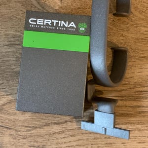 Może przedstawiać: Szare pudełko na zegarek z napisem "CERTINA SWISS WATCHES SINCE 1888" i zielonym logo z literami "DS". Pudełko leży na drewnianej powierzchni obok szarego paska do zegarka.