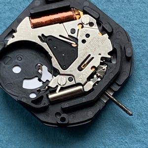 May include: A quartz watch movement mechanism featuring a black plastic base, a silver metal plate with the text "ONE JEWEL ORIENT WATCH CO. LTD. JAPAN" and "HT710" engraved, a copper wire coil, and a thin metal stem extending from the side.
