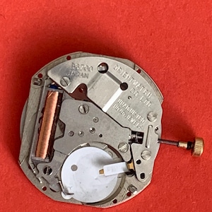 K&ouml;nnte beinhalten: Nahaufnahme eines Uhrwerks mit dem Text "B6700 JAPAN" und "ORIENT WATCH CO. LTD.". Das Uhrwerk ist silberfarben mit einer goldfarbenen Krone und einer kupferfarbenen Komponente. Der Hintergrund ist rot.