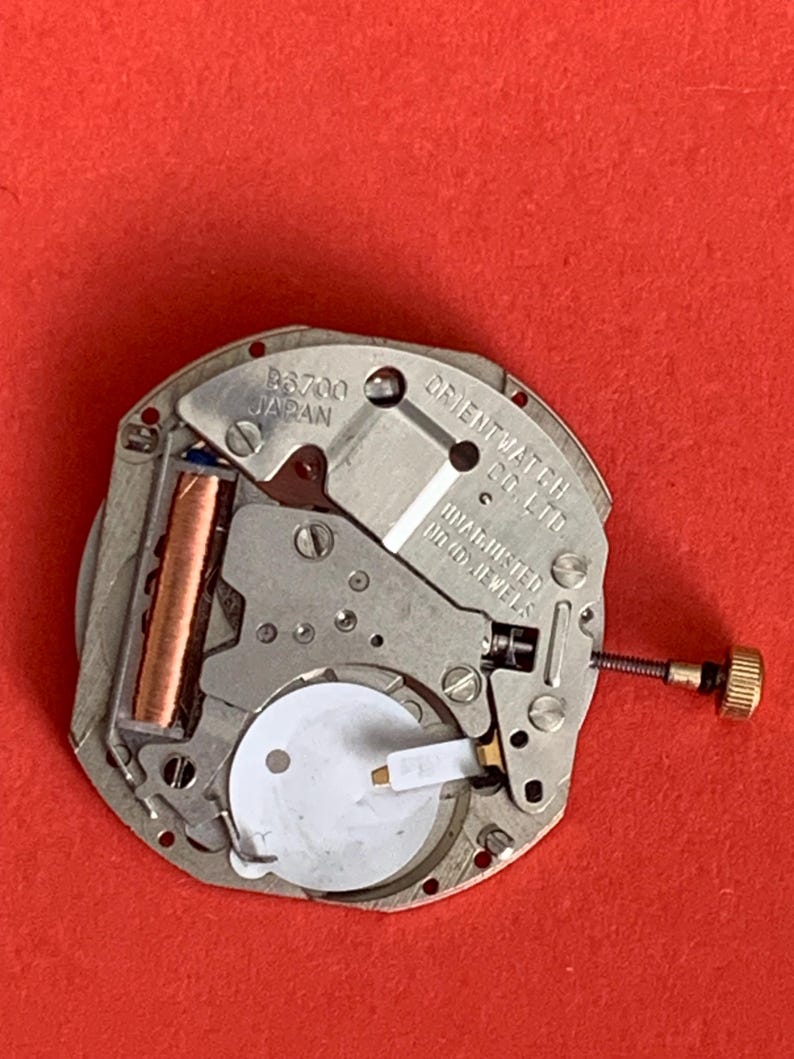 K&ouml;nnte beinhalten: Nahaufnahme eines Uhrwerks mit dem Text "B6700 JAPAN" und "ORIENT WATCH CO. LTD.". Das Uhrwerk ist silberfarben mit einer goldfarbenen Krone und einer kupferfarbenen Komponente. Der Hintergrund ist rot.