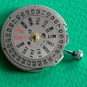 May include: A silver watch movement with a day and date dial. The dial displays the days of the week in English and numbers 1-31. A small crown is visible on the right side. The background is a solid green.