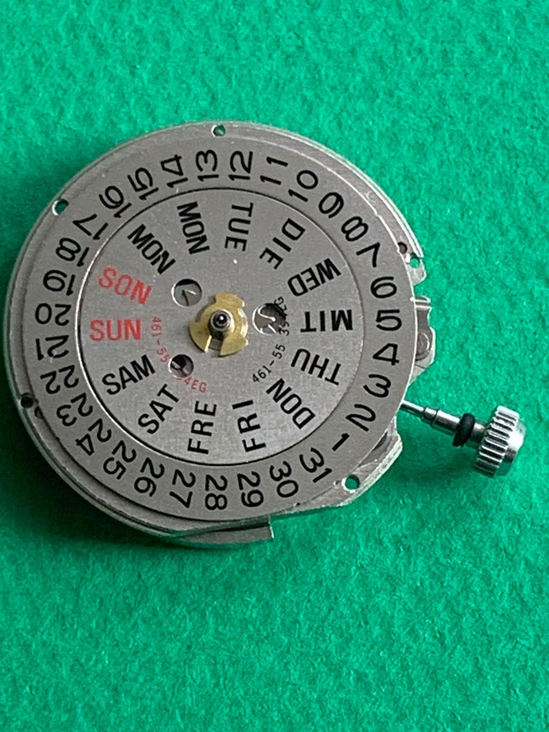 May include: A silver watch movement with a day and date dial. The dial displays the days of the week in English and numbers 1-31. A small crown is visible on the right side. The background is a solid green.