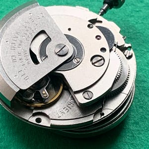 May include: Close-up of a watch movement with intricate gears, screws, and a winding stem. The metal components are silver and gold, with the text "ORIENT" and "ORIENT WATCH CO. LTD. JAPAN" visible. The background is a solid green.