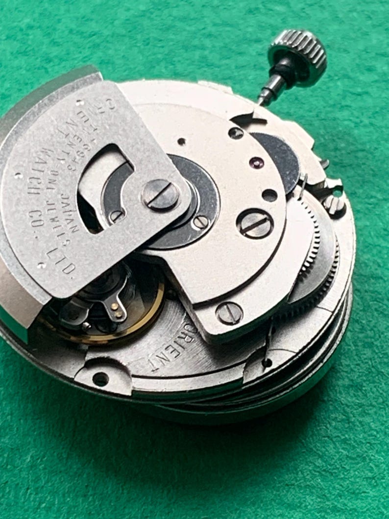 May include: Close-up of a watch movement with intricate gears, screws, and a winding stem. The metal components are silver and gold, with the text "ORIENT" and "ORIENT WATCH CO. LTD. JAPAN" visible. The background is a solid green.