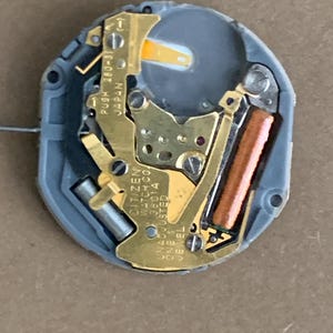 May include: A close-up of a Citizen watch movement. The movement features gold-colored metal components, a copper-colored coil, and a gray plastic casing. Text on the movement includes "CITIZEN WATCH CO. 38014 UNADJUSTED ONE JEWEL C" and "PUSH 280-31 JAPAN".