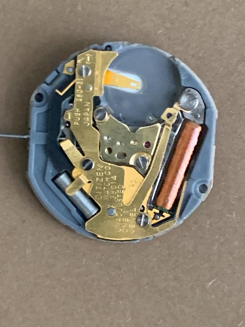 May include: A close-up of a Citizen watch movement. The movement features gold-colored metal components, a copper-colored coil, and a gray plastic casing. Text on the movement includes "CITIZEN WATCH CO. 38014 UNADJUSTED ONE JEWEL C" and "PUSH 280-31 JAPAN".