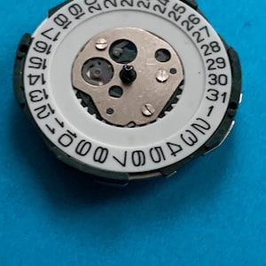May include: A watch movement component with a white dial and a black outer ring. The dial displays numbers 1-31, indicating the days of the month. The centre features a silver-coloured mechanical mechanism.