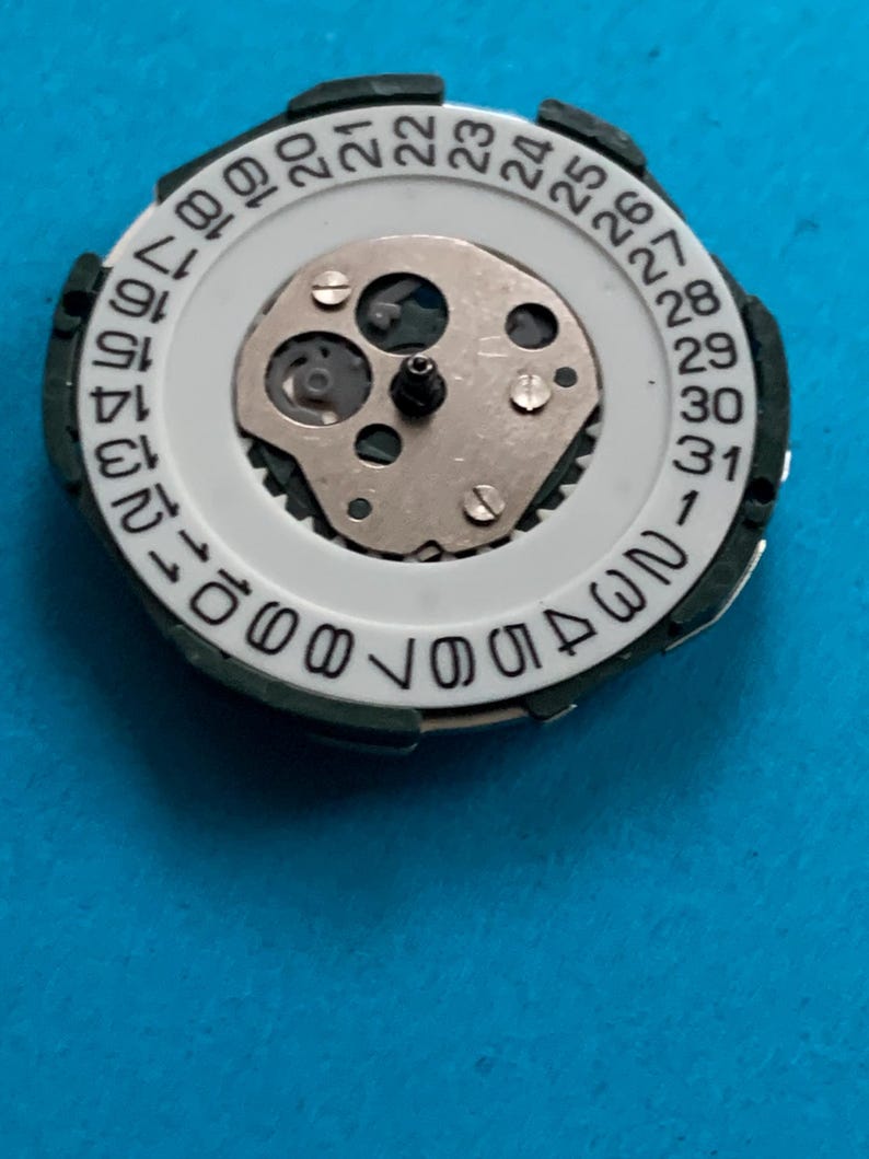 May include: A watch movement component with a white dial and a black outer ring. The dial displays numbers 1-31, indicating the days of the month. The centre features a silver-coloured mechanical mechanism.