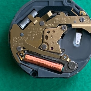 May include: Close-up of a Citizen watch movement. The watch mechanism features gold-colored gears, a copper coil, and a silver battery compartment. The text "CITIZEN WATCH CO. 3810A JAPAN" is visible.