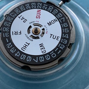 May include: A detailed view of a watch movement with a white face, displaying black numbers and day indicators. The days of the week are in black and red. The dial is encased in silver metal, with a crown at the top.