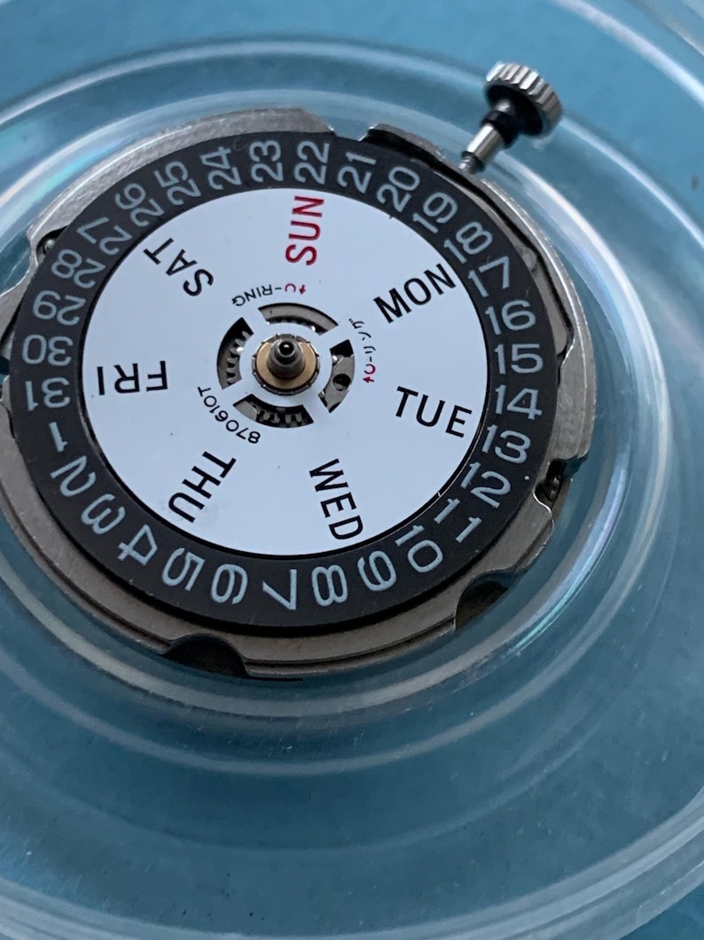 May include: A detailed view of a watch movement with a white face, displaying black numbers and day indicators. The days of the week are in black and red. The dial is encased in silver metal, with a crown at the top.