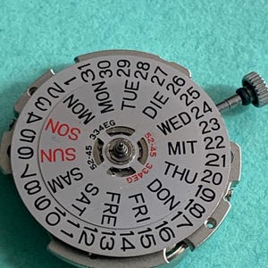 May include: A silver watch movement with a day and date display. The dial features the days of the week in English, German, and other languages, along with numbers for the date. The text "334EG" and "52-45" are also visible.