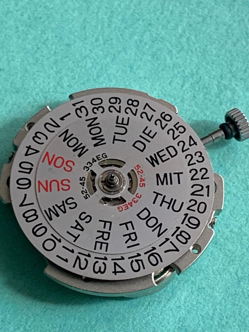 May include: A silver watch movement with a day and date display. The dial features the days of the week in English, German, and other languages, along with numbers for the date. The text "334EG" and "52-45" are also visible.