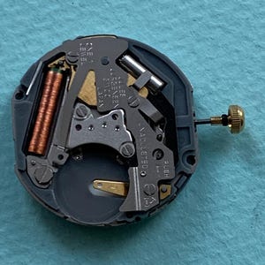 K&ouml;nnte beinhalten: Nahaufnahme eines Uhrwerks mit komplexen mechanischen Komponenten. Der Mechanismus zeigt eine Kupferspule, silberfarbene Metallteile und eine goldfarbene Krone. Der Text "Jules J&uuml;rgensen 21100 Japan" ist sichtbar.