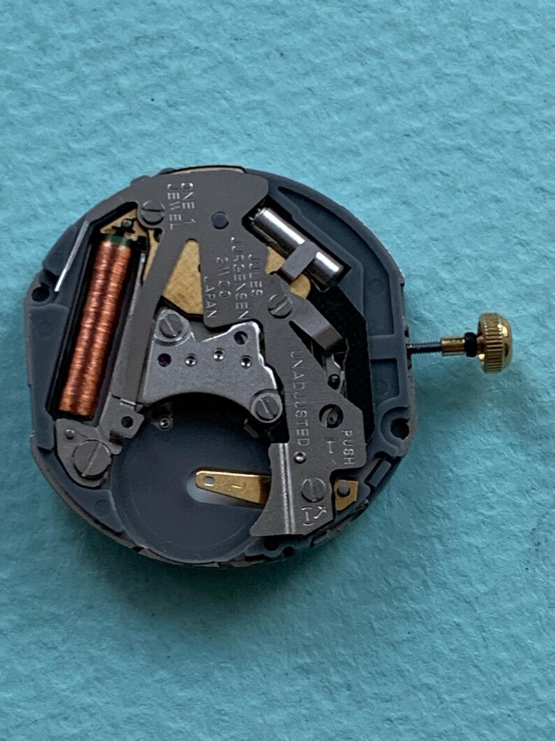 K&ouml;nnte beinhalten: Nahaufnahme eines Uhrwerks mit komplexen mechanischen Komponenten. Der Mechanismus zeigt eine Kupferspule, silberfarbene Metallteile und eine goldfarbene Krone. Der Text "Jules J&uuml;rgensen 21100 Japan" ist sichtbar.
