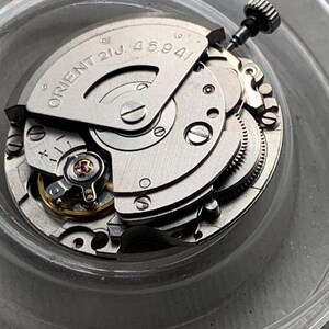 Pu&ograve; includere: Primo piano di un movimento di orologio argentato con la scritta "ORIENT 21J. 46941". L'intricato meccanismo presenta ingranaggi, viti e una corona. Il movimento dell'orologio &egrave; in un contenitore di plastica trasparente.
