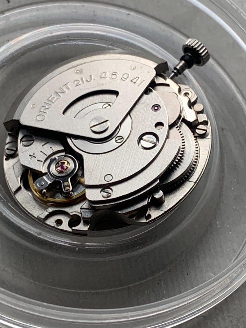 Pu&ograve; includere: Primo piano di un movimento di orologio argentato con la scritta "ORIENT 21J. 46941". L'intricato meccanismo presenta ingranaggi, viti e una corona. Il movimento dell'orologio &egrave; in un contenitore di plastica trasparente.