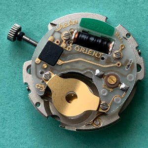 May include: A close-up of a watch movement with the word "JAPAN" and "ORIENT" visible. The movement has a silver-colored base with gold and black components, including a black coil and a green piece.