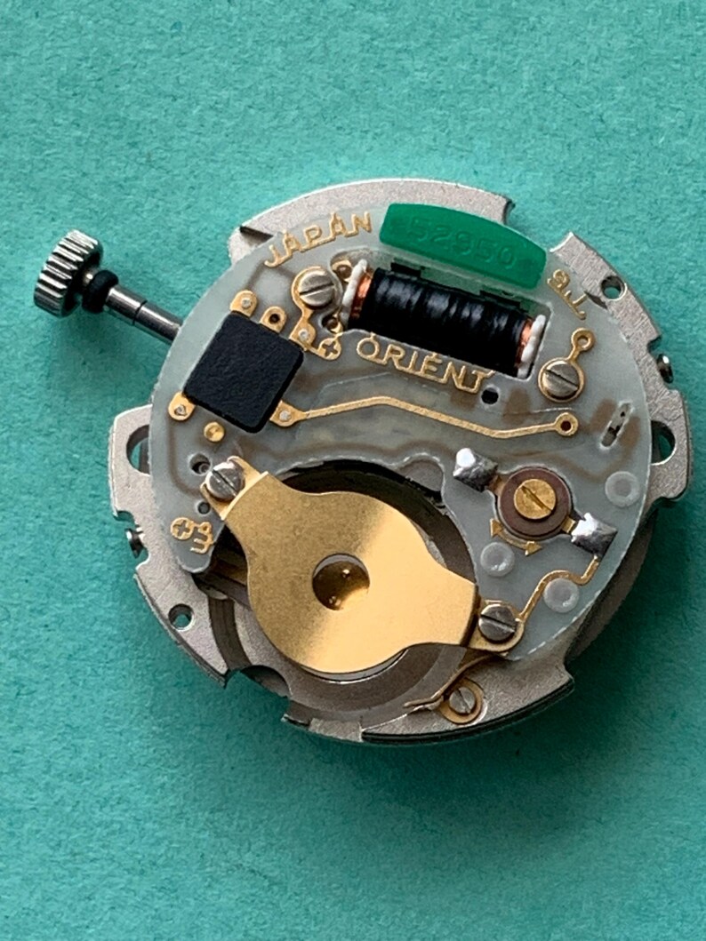 May include: A close-up of a watch movement with the word "JAPAN" and "ORIENT" visible. The movement has a silver-colored base with gold and black components, including a black coil and a green piece.