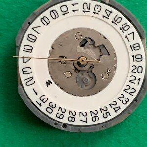 May include: Close-up of a watch movement with a white dial and a gray outer ring. The dial features black numbers indicating the days of the month. A gold-colored hand points to the current date. The watch mechanism is visible.