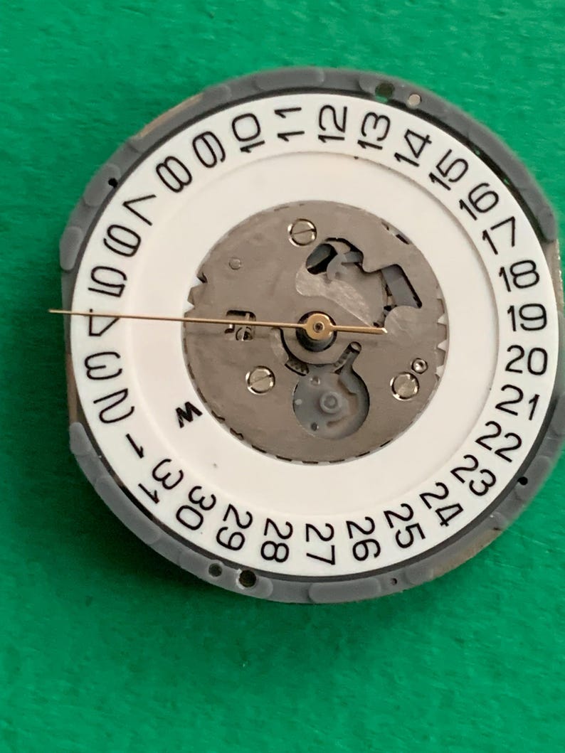 May include: Close-up of a watch movement with a white dial and a gray outer ring. The dial features black numbers indicating the days of the month. A gold-colored hand points to the current date. The watch mechanism is visible.