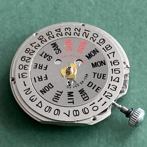 May include: A silver watch movement with a day and date dial. The dial shows the days of the week in English, German, and other languages, and the date numbers. A small crown is visible on the side.