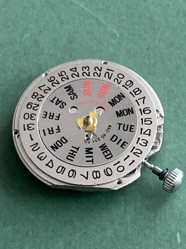 May include: A silver watch movement with a day and date dial. The dial shows the days of the week in English, German, and other languages, and the date numbers. A small crown is visible on the side.