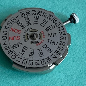 May include: A silver watch movement with a day and date display. The dial features the days of the week in English, German, and other languages, along with numbers for the date. A small crown is visible on the side.