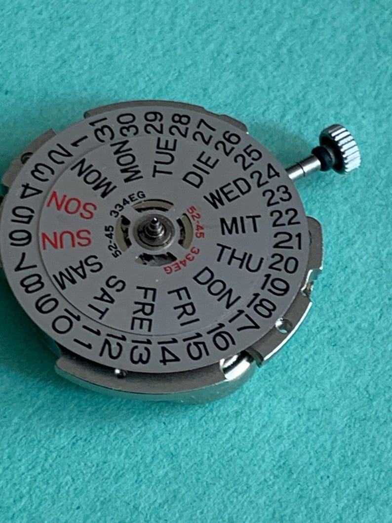 May include: A silver watch movement with a day and date display. The dial features the days of the week in English, German, and other languages, along with numbers for the date. A small crown is visible on the side.