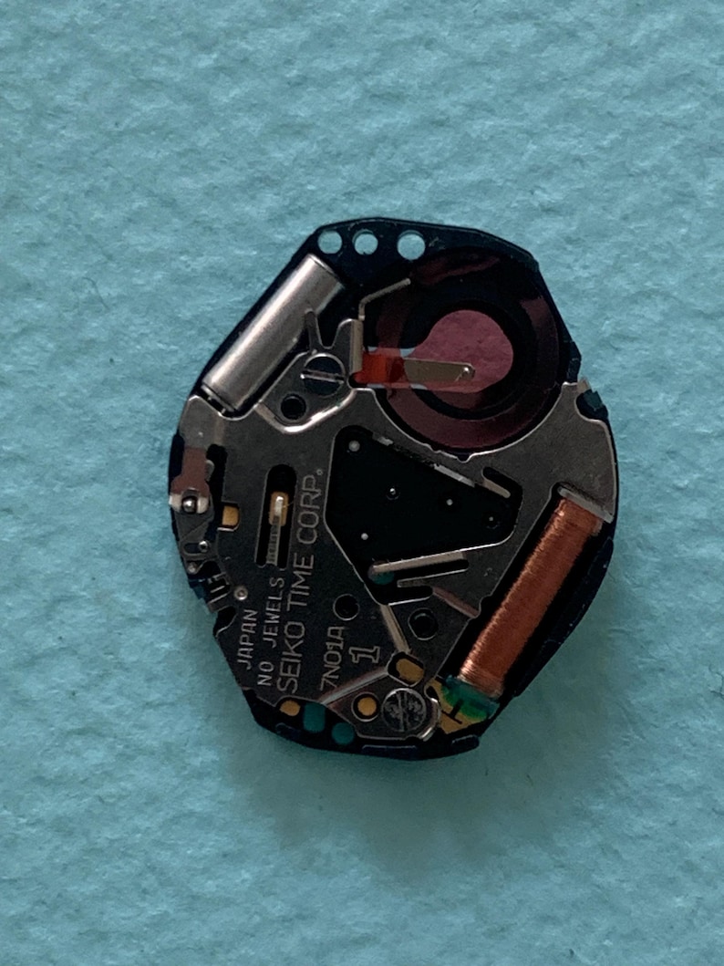 May include: A close-up of a watch movement with the text "JAPAN NO JEWELS SEIKO TIME CORP". The movement has a black base with silver and copper components. The watch movement is a 7N01A model.