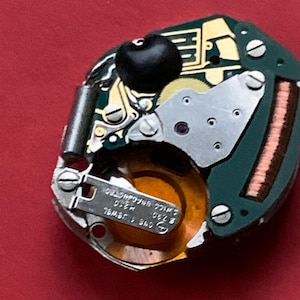 May include: Close-up of a watch movement with a green circuit board, gold-colored gears, and silver screws. A black component is visible at the top. The watch movement has the text "SWISS UNADJUSTED" and "ONE 1 JEWEL".