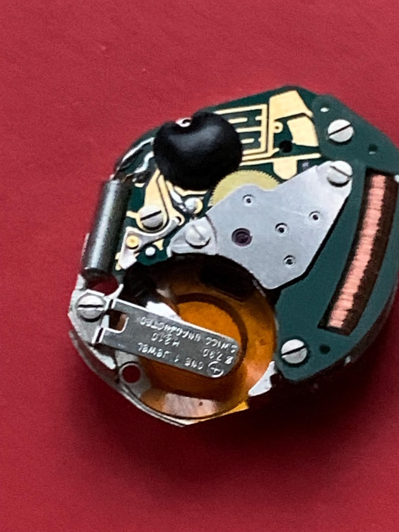 May include: Close-up of a watch movement with a green circuit board, gold-colored gears, and silver screws. A black component is visible at the top. The watch movement has the text "SWISS UNADJUSTED" and "ONE 1 JEWEL".