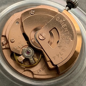 Puede incluir: Primer plano de un movimiento de reloj con engranajes y componentes dorados. El texto "ORIENT 21 JEWELS JAPAN 46744" es visible en una placa met&aacute;lica. El mecanismo del reloj es intrincado y detallado.
