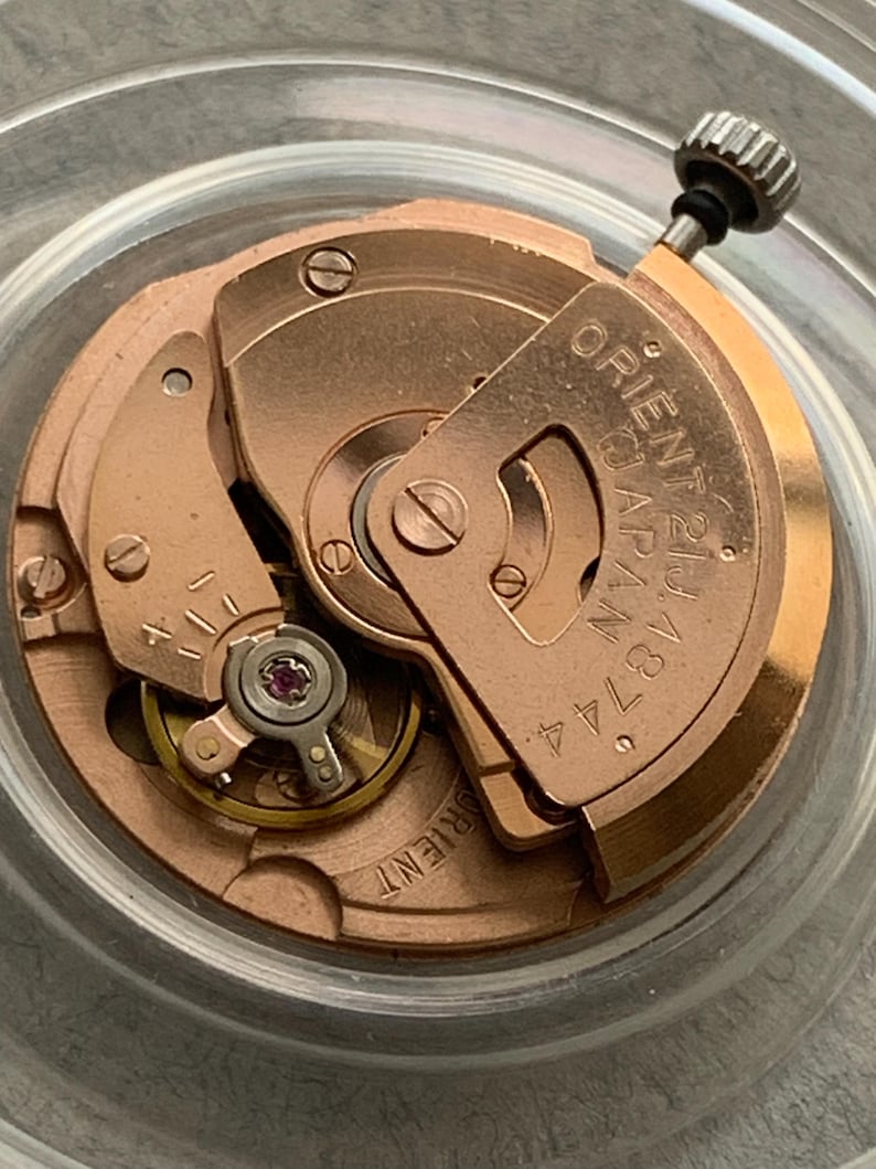 Puede incluir: Primer plano de un movimiento de reloj con engranajes y componentes dorados. El texto "ORIENT 21 JEWELS JAPAN 46744" es visible en una placa met&aacute;lica. El mecanismo del reloj es intrincado y detallado.