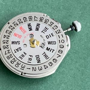 May include: A silver watch movement with a white dial showing the days of the week in English and numbers 1-31. The days are abbreviated, and the word "SUN" is in red. A small crown is visible on the right side.
