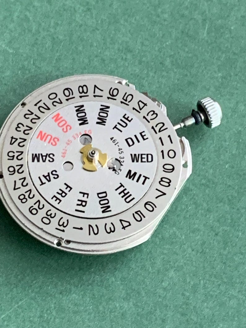 May include: A silver watch movement with a white dial showing the days of the week in English and numbers 1-31. The days are abbreviated, and the word "SUN" is in red. A small crown is visible on the right side.