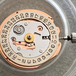 Puede incluir: Primer plano de un movimiento de reloj con una esfera circular que muestra los n&uacute;meros del 1 al 31. El mecanismo tiene una combinaci&oacute;n de colores plateado y dorado, con peque&ntilde;os engranajes, tornillos y una joya. Una peque&ntilde;a herramienta est&aacute; adjunta al lado.