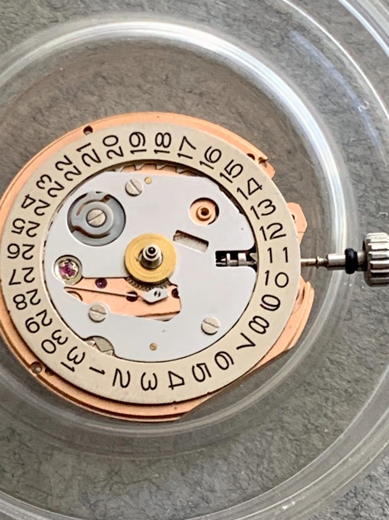Puede incluir: Primer plano de un movimiento de reloj con una esfera circular que muestra los n&uacute;meros del 1 al 31. El mecanismo tiene una combinaci&oacute;n de colores plateado y dorado, con peque&ntilde;os engranajes, tornillos y una joya. Una peque&ntilde;a herramienta est&aacute; adjunta al lado.