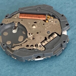 May include: A close-up of a watch movement, featuring intricate metal components and a copper-colored coil. The movement is encased in a gray plastic frame. The image is taken from a top-down perspective, showcasing the internal mechanisms of the watch.