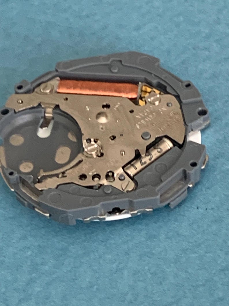 May include: A close-up of a watch movement, featuring intricate metal components and a copper-colored coil. The movement is encased in a gray plastic frame. The image is taken from a top-down perspective, showcasing the internal mechanisms of the watch.