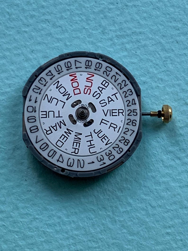K&ouml;nnte beinhalten: Nahaufnahme eines Uhrwerks mit wei&szlig;em Zifferblatt, das die Wochentage und Daten anzeigt. Das Zifferblatt ist von einem schwarzen Ring umgeben. Eine kleine goldfarbene Krone ist an der Seite angebracht.