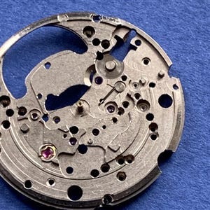 Puede incluir: Primer plano de un movimiento de reloj plateado con intrincados engranajes, tornillos y joyas. Los componentes mec&aacute;nicos est&aacute;n dispuestos en un patr&oacute;n circular, mostrando la precisi&oacute;n del proceso de fabricaci&oacute;n de relojes. Fondo azul liso.