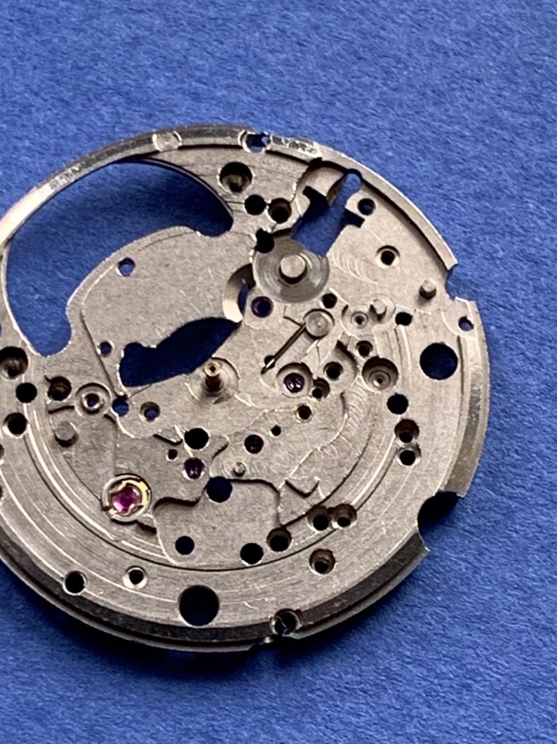 Puede incluir: Primer plano de un movimiento de reloj plateado con intrincados engranajes, tornillos y joyas. Los componentes mec&aacute;nicos est&aacute;n dispuestos en un patr&oacute;n circular, mostrando la precisi&oacute;n del proceso de fabricaci&oacute;n de relojes. Fondo azul liso.