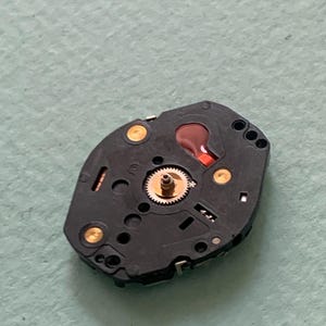 May include: A close-up of a black watch component featuring a small, gold-coloured gear at its centre. The component has several gold-coloured contact points and a red liquid-filled reservoir. The background is a light green surface.