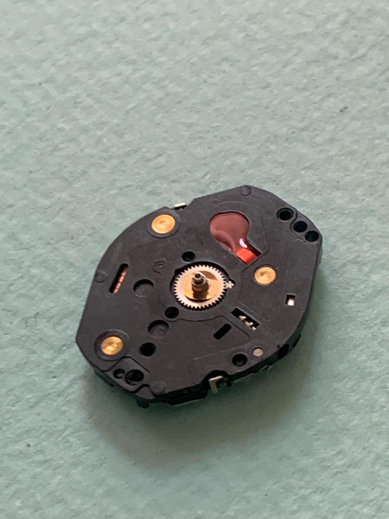 May include: A close-up of a black watch component featuring a small, gold-coloured gear at its centre. The component has several gold-coloured contact points and a red liquid-filled reservoir. The background is a light green surface.