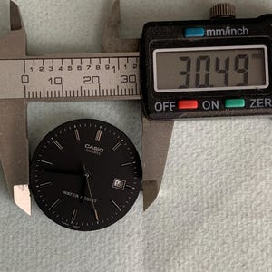 Puede incluir: Una esfera de reloj Casio Quartz negra con manecillas y marcadores blancos, y la fecha 14. La esfera del reloj se mide con un calibrador digital, mostrando una medida de 30,49 mm.