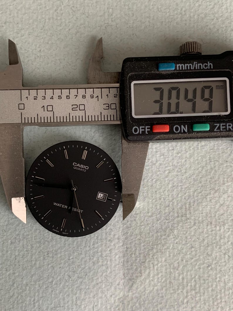 Puede incluir: Una esfera de reloj Casio Quartz negra con manecillas y marcadores blancos, y la fecha 14. La esfera del reloj se mide con un calibrador digital, mostrando una medida de 30,49 mm.